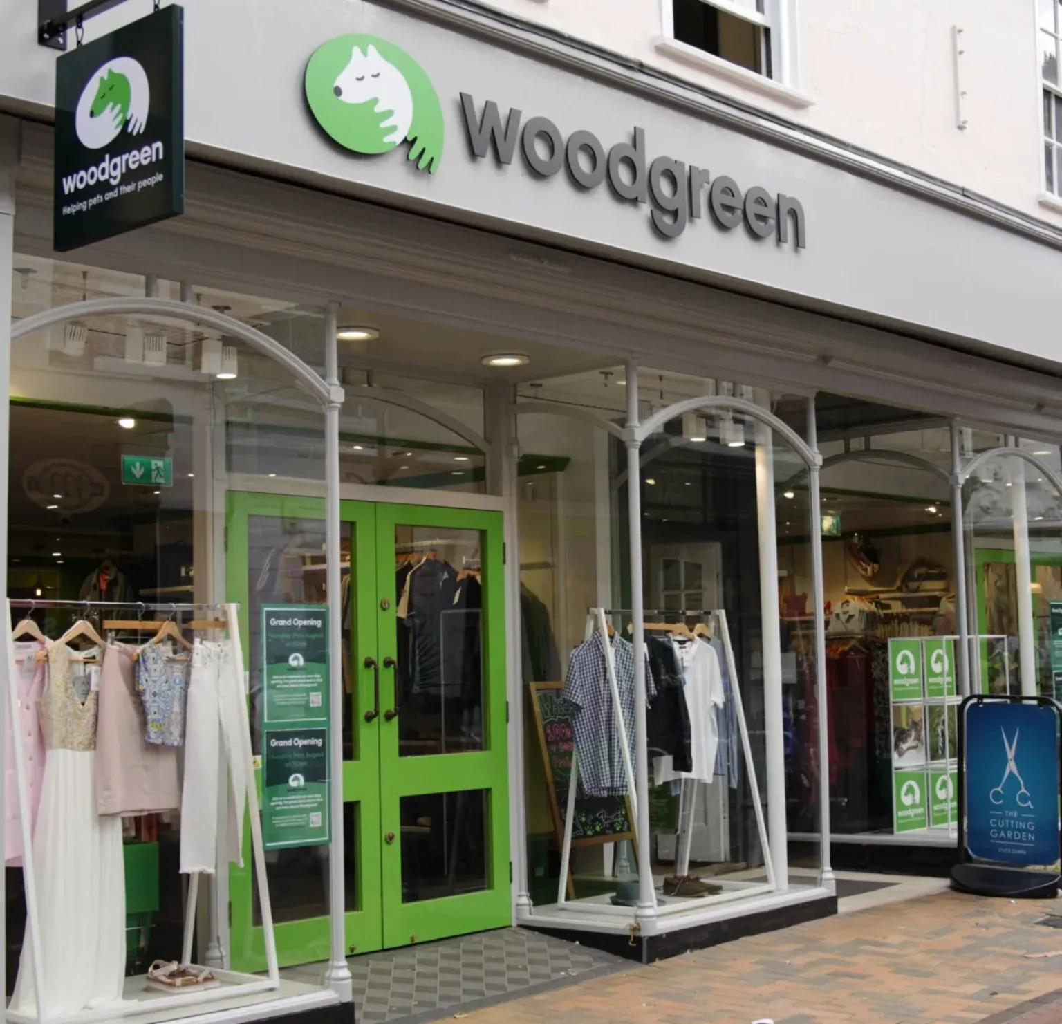 Woodgreen charity shop exterior with green doors, large windows displaying clothes on mannequins, and a sign above the entrance featuring a white dog logo on a green background. A blue donation bin stands outside.