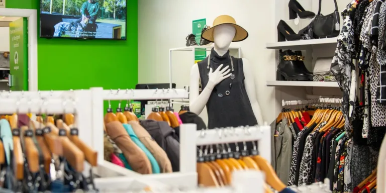A clothing store display with a mannequin wearing a sleeveless black dress and straw hat, surrounded by racks of colorful clothes. Shelves with shoes are visible, and a TV screen hangs on a bright green wall in the background.