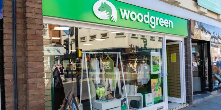 Shopfront of the St Neots charity shop by Woodgreen, with a green sign featuring a white dog logo. Display window shows clothes, accessories, and decor items.