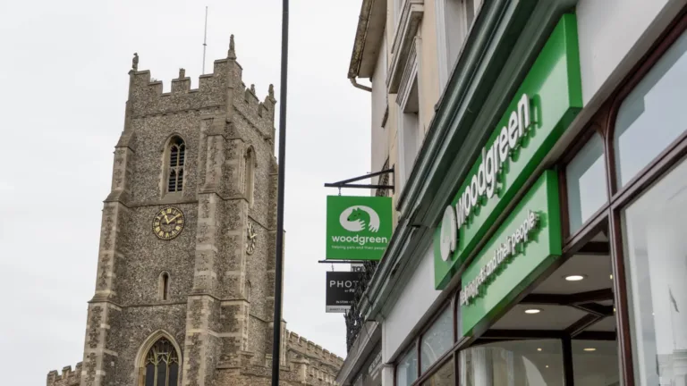 A stone church tower with a clock stands next to a street lined with shops, including a Sudbury charity shop with bright green signs reading woodgreen and a logo featuring an animal.