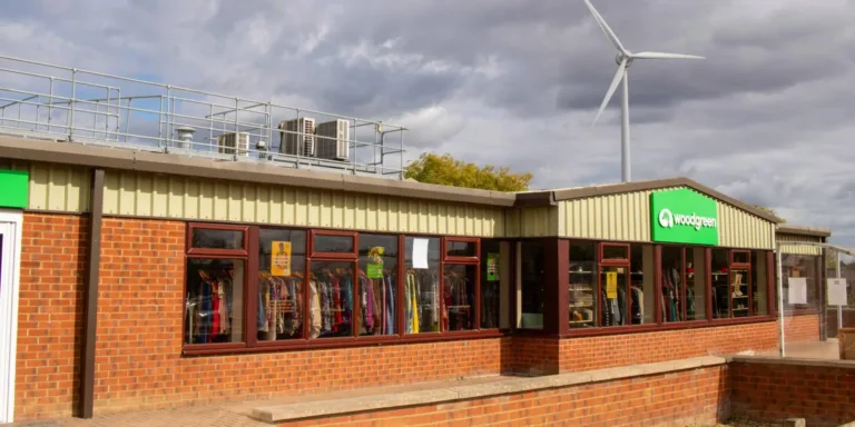 A single-story brick building with large windows displays clothing inside. The green “Woodgreen” sign is visible on the wall, marking this as a Godmanchester charity shop. A wind turbine and cloudy sky are in the background.
