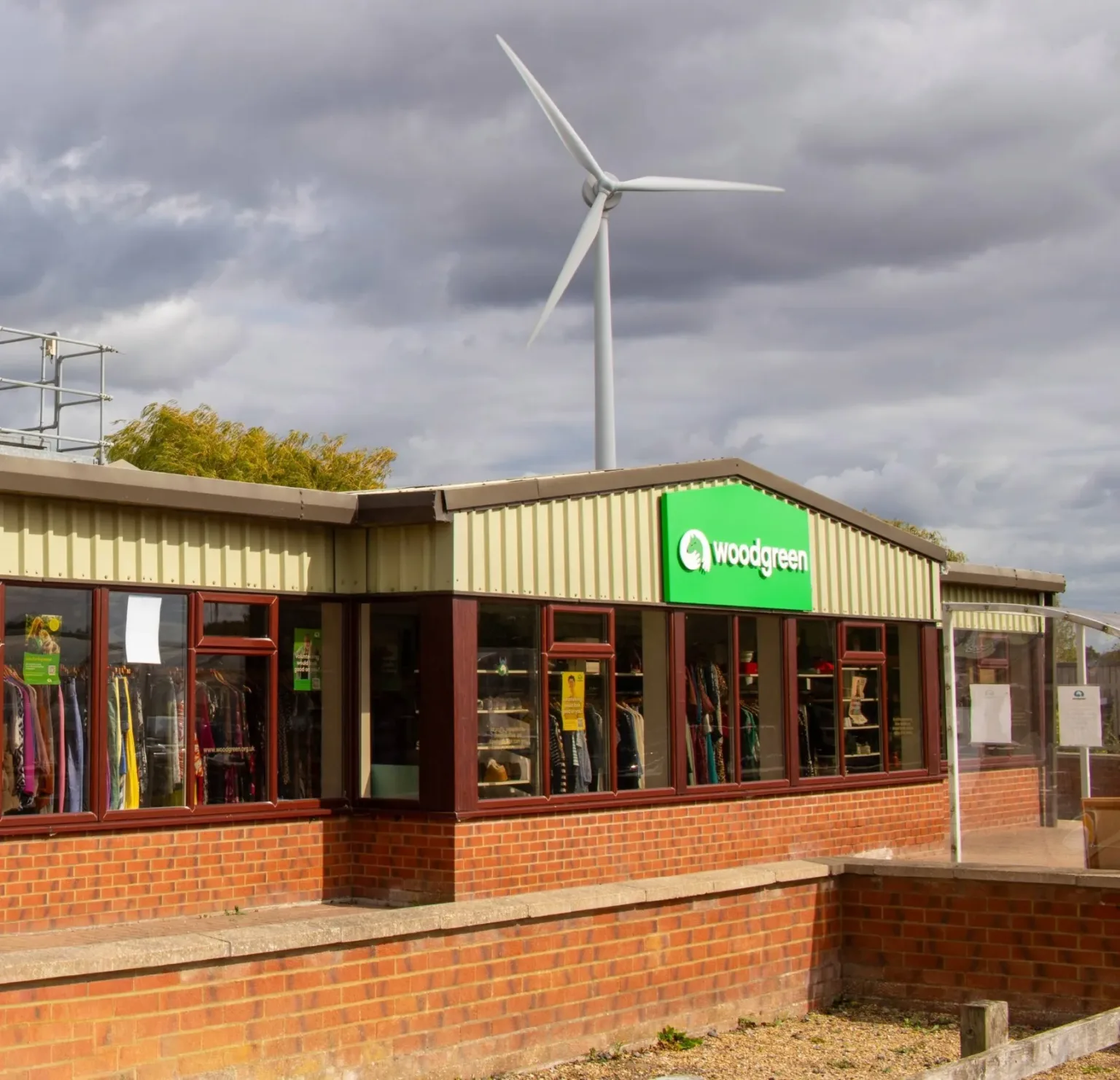 A Godmanchester charity shop with a green woodgreen sign displays clothes and goods in the windows. A large wind turbine stands behind the building under a cloudy sky.