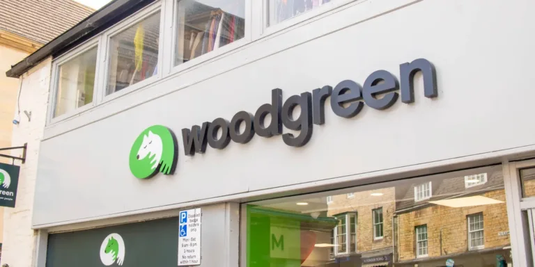 A storefront with a sign that reads woodgreen in black letters, next to a green circle logo featuring a white animal, possibly a dog or cat, on Stamford’s busy street with buildings reflected in the windows.