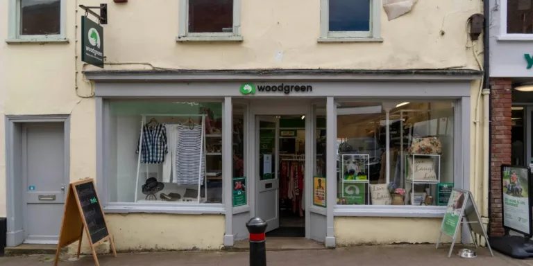 A charity shop named Woodgreen in Woodbridge features a glass storefront displaying clothes and accessories. Signs and posters are visible outside and inside the shop, and the entrance door is open.