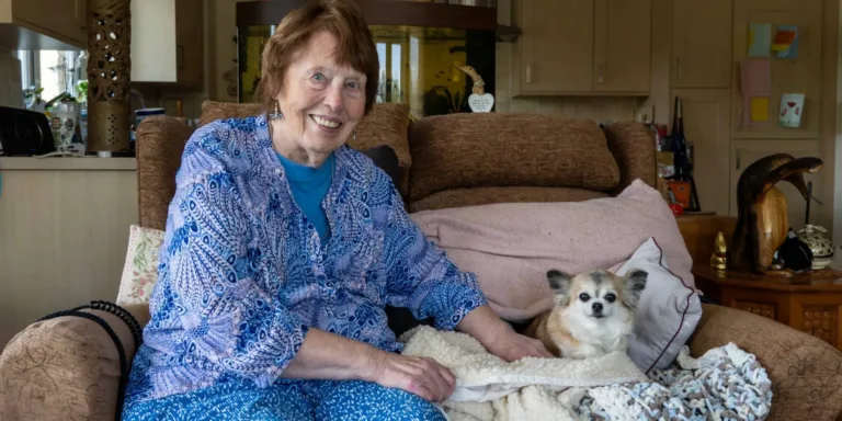 An elderly woman with short brown hair, wearing a blue patterned outfit, sits smiling on a cushy chair next to a small fluffy dog wrapped in a blanket. They are in a cozy living room with wood cabinets and decor in the background.