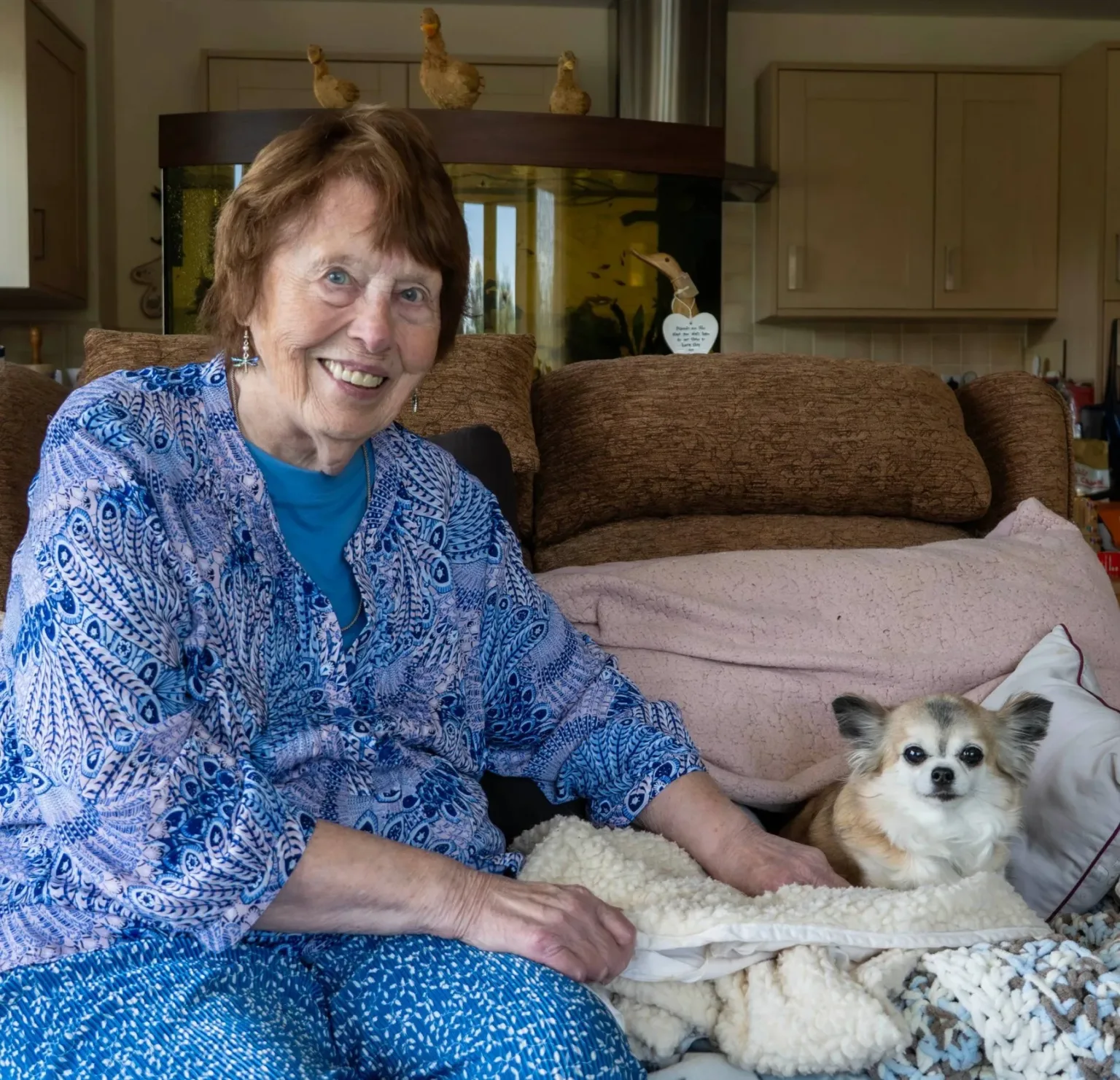 An elderly woman with short brown hair and a blue patterned blouse sits on a couch, smiling, next to a small fluffy dog lying on a blanket. Cozy home environment with cabinets and a fish tank in the background.