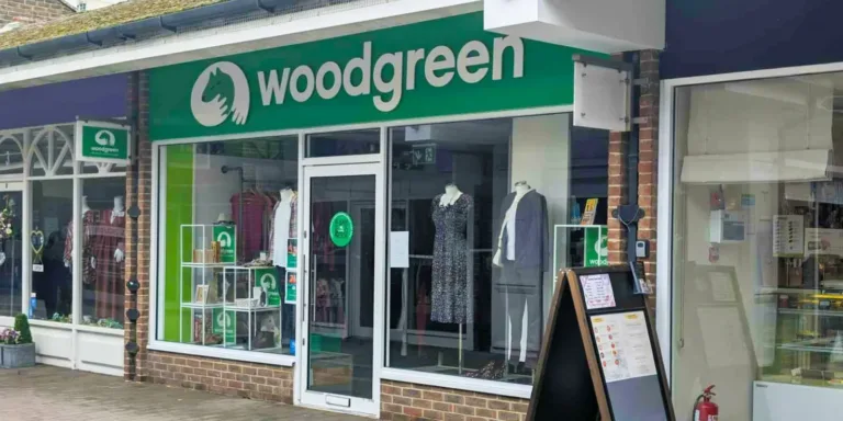 A shopfront in Bicester with a green sign reading woodgreen, displaying clothing and accessories in the windows. The entrance features glass doors, and there's a sidewalk sandwich board outside.
