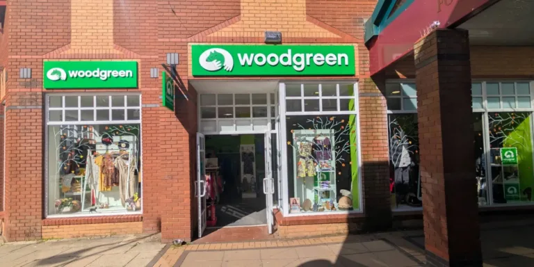 A Woodgreen charity shop in Daventry with a bright green sign above the entrance. The shop window displays clothing and various items inside. The sunlit brick building has a paved walkway in front.