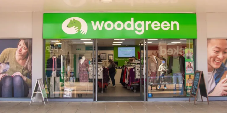 A shopfront in Dunstable with a bright green sign reading woodgreen and a logo of a hand holding an animal. Inside, clothing racks are visible, and posters with people and pets decorate the entrance.