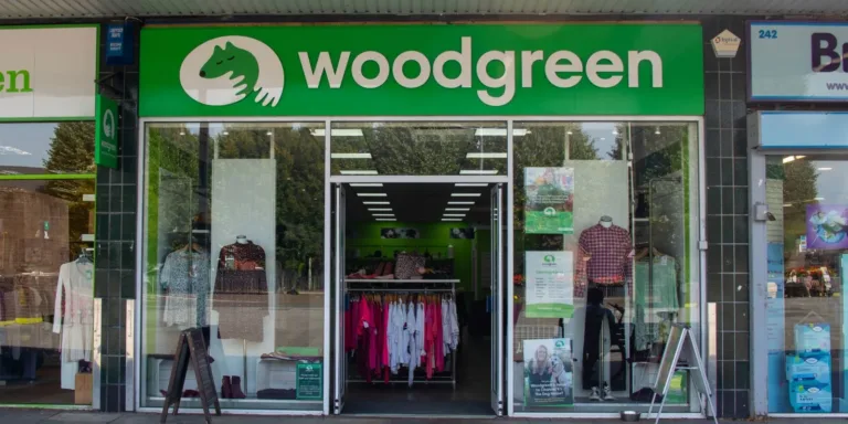 The image shows the storefront of a Woodgreen Kempston charity shop with a green sign above the entrance. The shop displays clothing inside and promotional posters in the front windows.