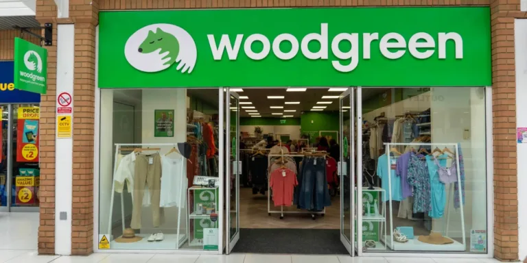 The entrance to the Newmarket charity shop, Woodgreen, features a bright green sign with a white logo of a hand and animal face. Inside, clothing racks display various garments, and open glass doors lead into the shopping mall.
