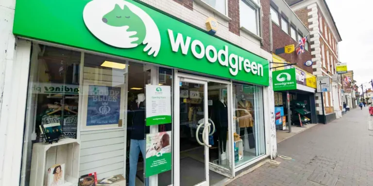 A green and white storefront for Woodgreen’s Newport Pagnell charity shop features large glass windows, clothing displays inside, and signs with a logo of hands cradling a pet. The shop sits on a brick-paved street alongside other stores.