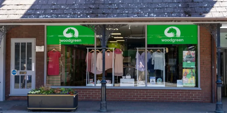 A Sandy charity shop with green woodgreen signs above large windows displays clothing on hangers. A brick planter with flowers sits on the sidewalk in front, and the store has a red brick exterior and a tiled roof.