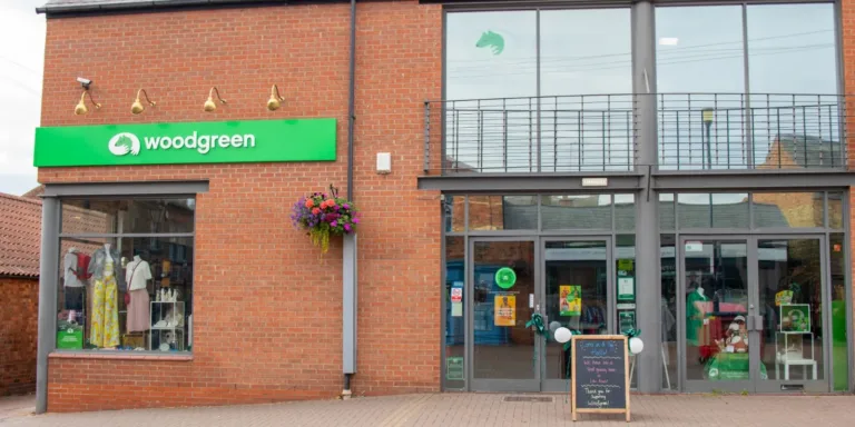 A brick building in Spalding with a green woodgreen sign, large glass entrance doors, a balcony, chalkboard sign outside, hanging flower basket, and a display window showcasing clothes and other items.