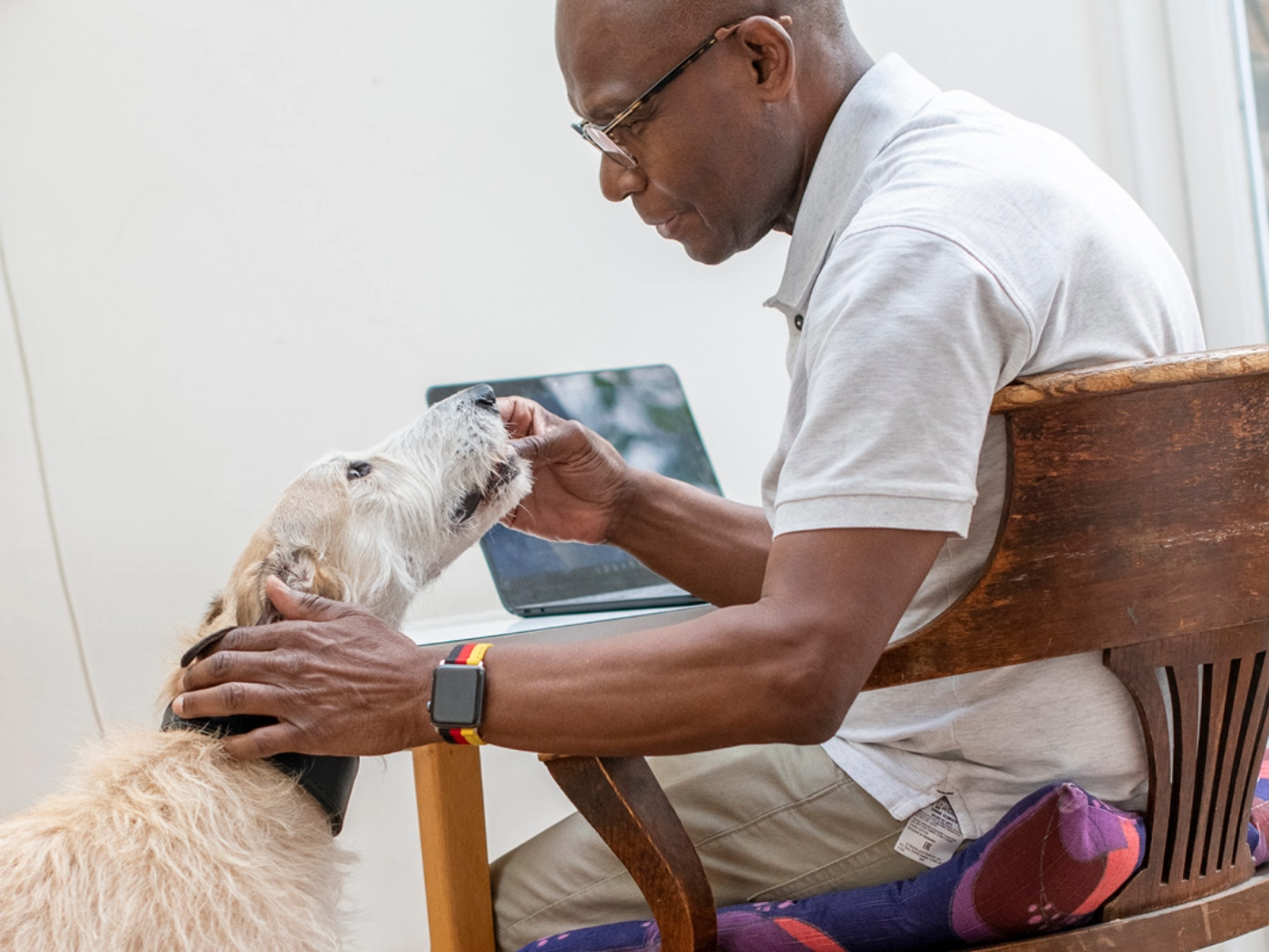 A man in glasses sits at a desk with a laptop, gently petting and feeding a light-colored, shaggy dog beside him. Wearing a smart watch and light polo, he shows the caring spirit found at Woodgreen pets charity.