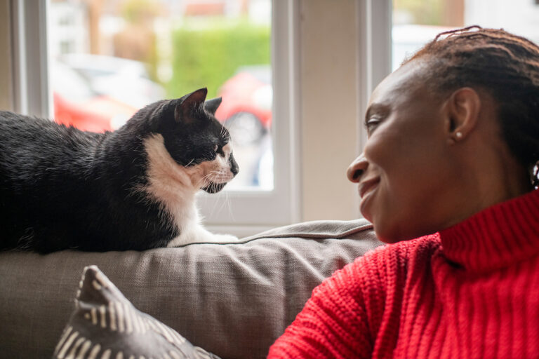 A woman in a red sweater smiles warmly at a black and white cat sitting on the back of a couch by a window.