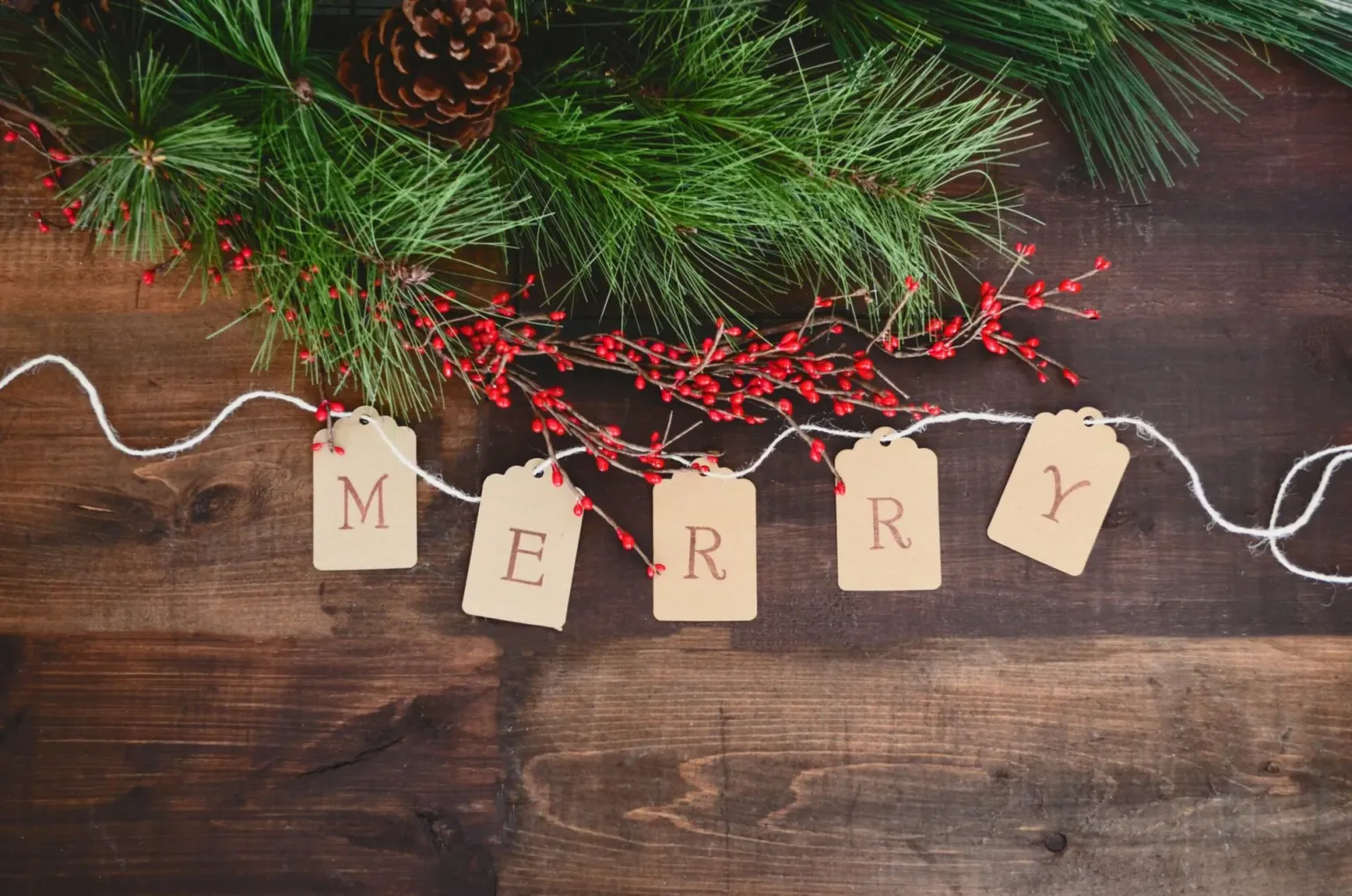 Top view of pine branches, red berries, and a pinecone on a wooden surface with string and tags spelling “MERRY” in red letters. Festive and rustic holiday decor.