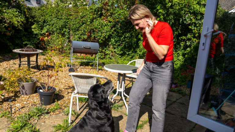 A woman in a red shirt smiles and waves at a black dog in a sunny backyard with patio furniture, plants, and a barbecue grill.