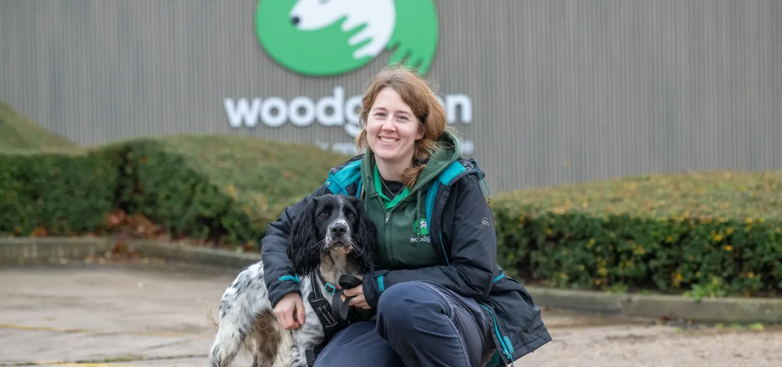 A woman in outdoor clothing kneels next to a black and white dog, both smiling at the camera, in front of a building with the wood part of a Woodgreen sign visible and greenery in the background.