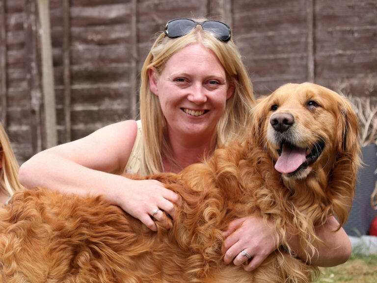 A woman with blonde hair and sunglasses on her head smiles while hugging a happy golden retriever outdoors, with a wooden fence in the background.