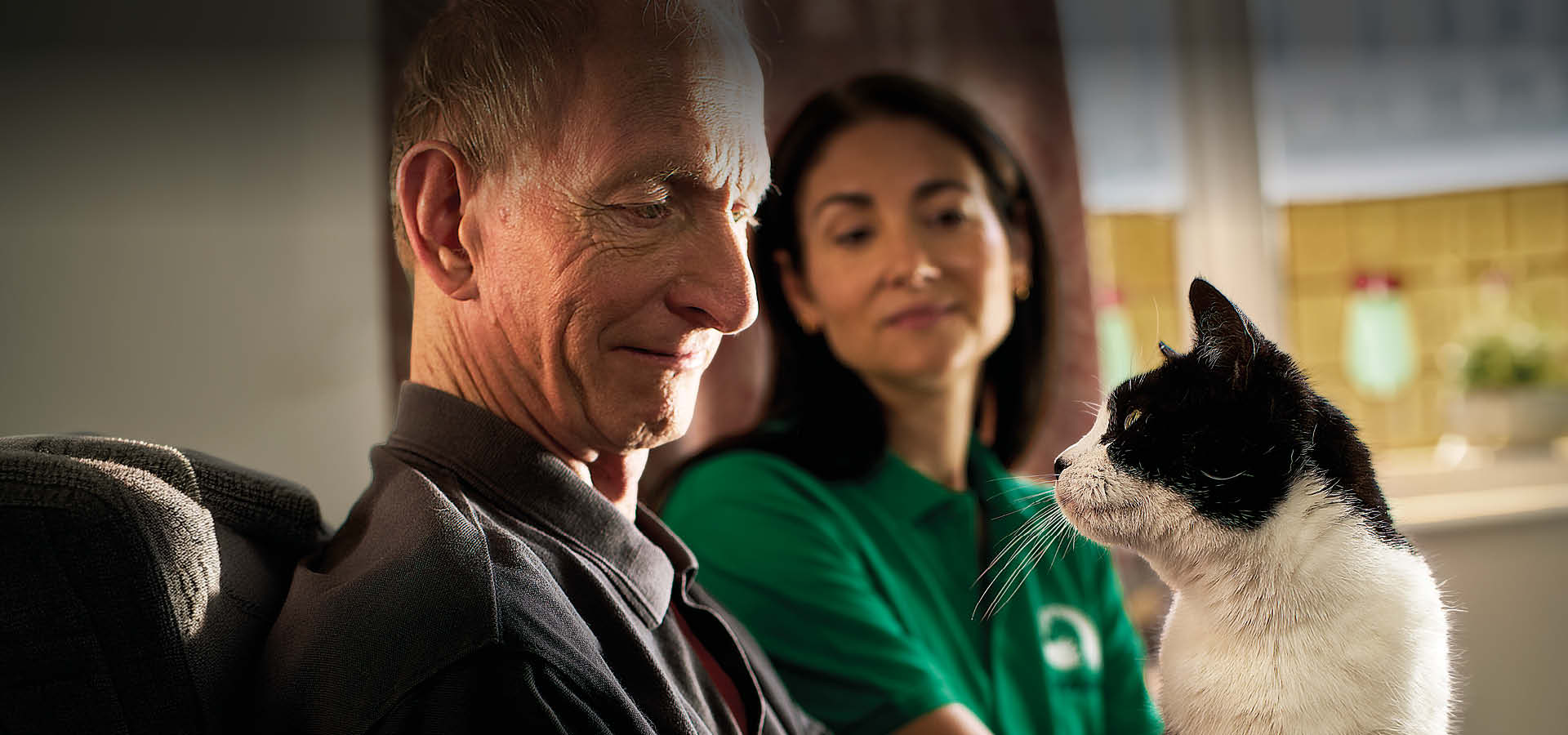 An elderly man smiles at a black and white cat on his lap, whilst a carer in a green shirt sits beside them, watching the heartwarming interaction—showcasing the joy that Woodgreen pets charity brings to people and their pets.
