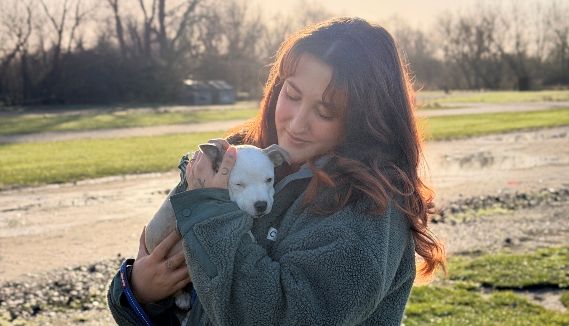 A woman with long brown hair gently holds a sleeping white puppy outdoors on a sunny day, with grass and trees in the background.