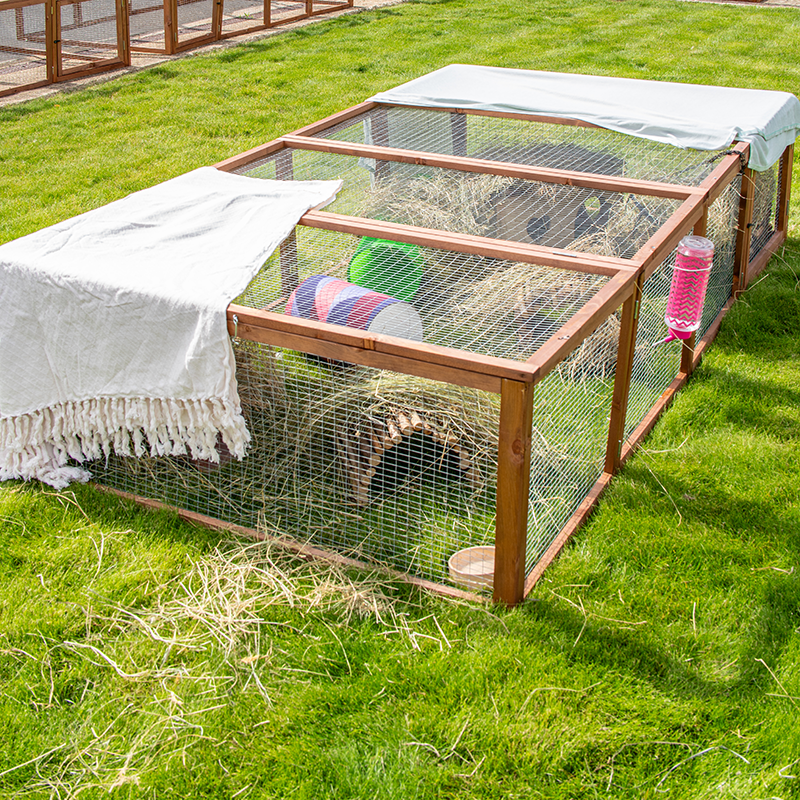 A wooden and wire outdoor enclosure sits on green grass, partly covered with a white fringed cloth. Inside are colourful toys, hay, and a water bottle, providing a play area for small animals like rabbits or guinea pigs.
