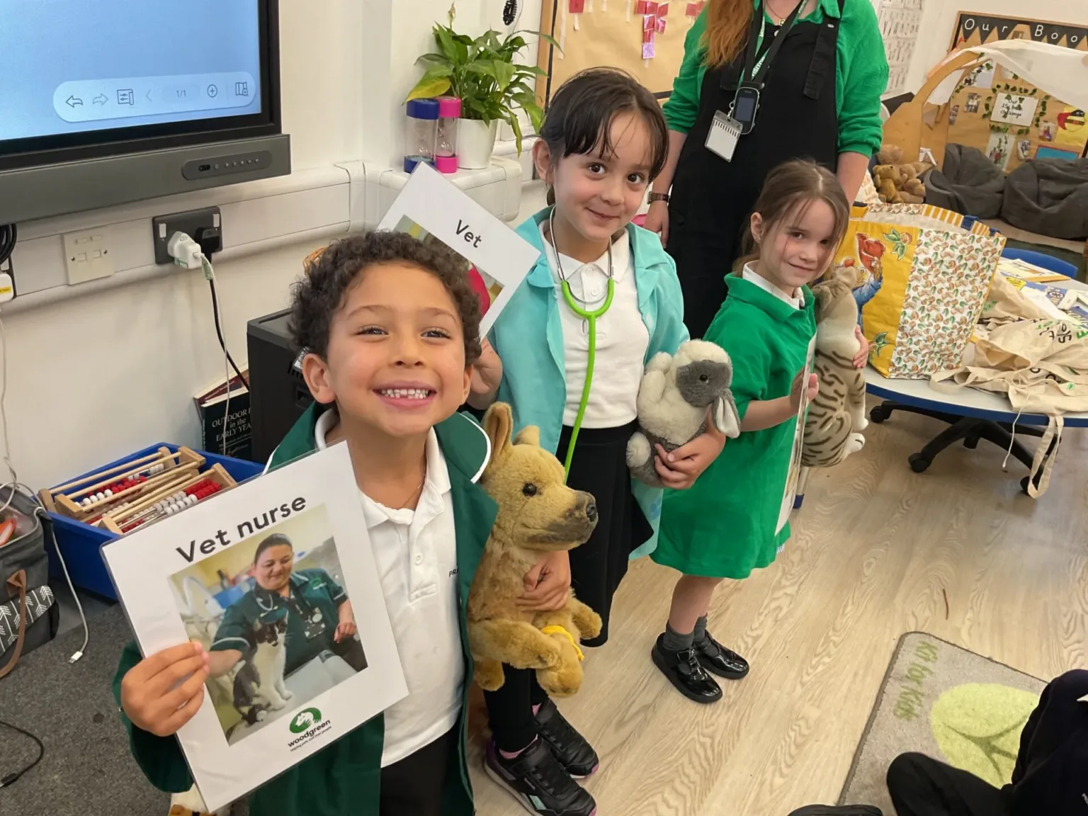 Three young children smile and hold soft toys and veterinary props in a classroom, with one child holding a paper labelled Vet nurse. A teacher stands behind them. The setting is colourful and playful.