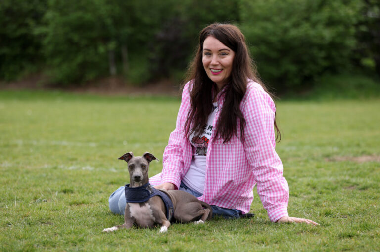 A woman in a pink chequered shirt sits on grass, smiling at the camera, with a small grey dog wearing a blue harness lying beside her. Trees and greenery are visible in the background.
