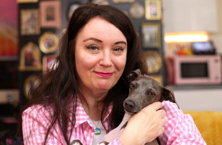 A woman with long dark hair in a pink checked shirt smiles while holding a small grey dog. The background shows framed photos and objects on a wall, and a pink microwave.
