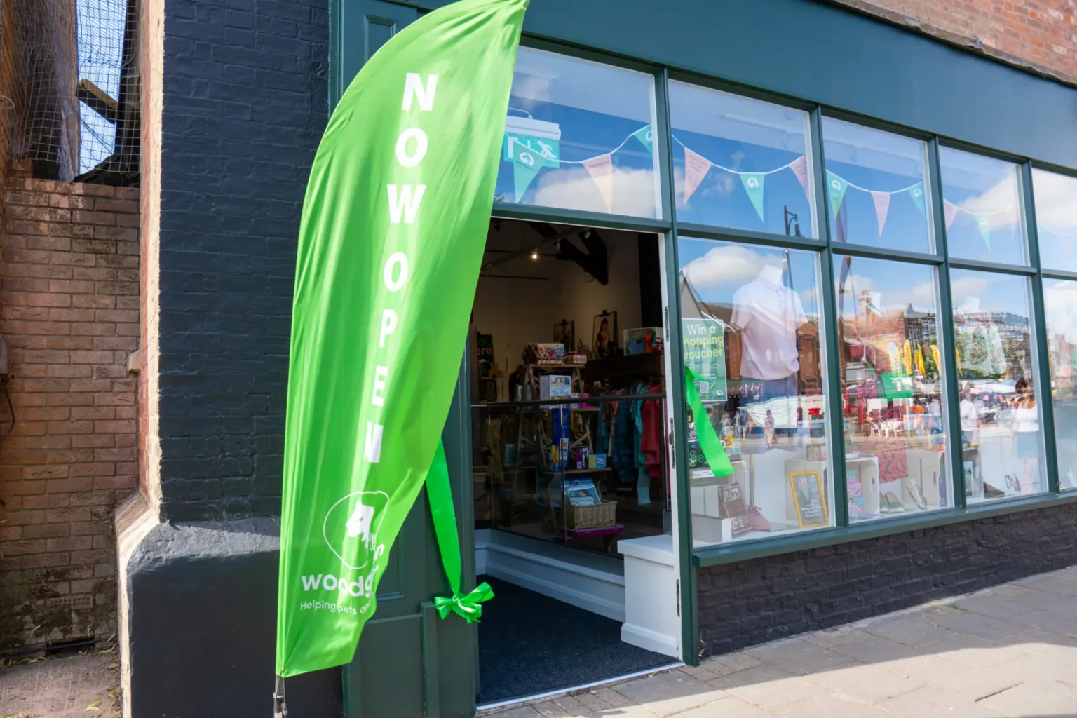 A bright green vertical flag reading NOW OPEN stands outside a shop with large windows displaying assorted colourful items and decorations, inviting customers inside.