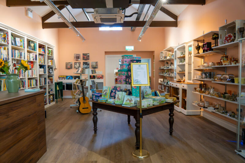 A well-lit gift shop with bookcases, display tables filled with gifts and books, a guitar, and various decorative items. A sign on a stand is positioned in front of the central table.
