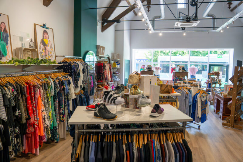 Clothing boutique interior with colourful dresses and shirts on rails, shoes displayed on a central table, and large windows letting in natural light from the street outside.