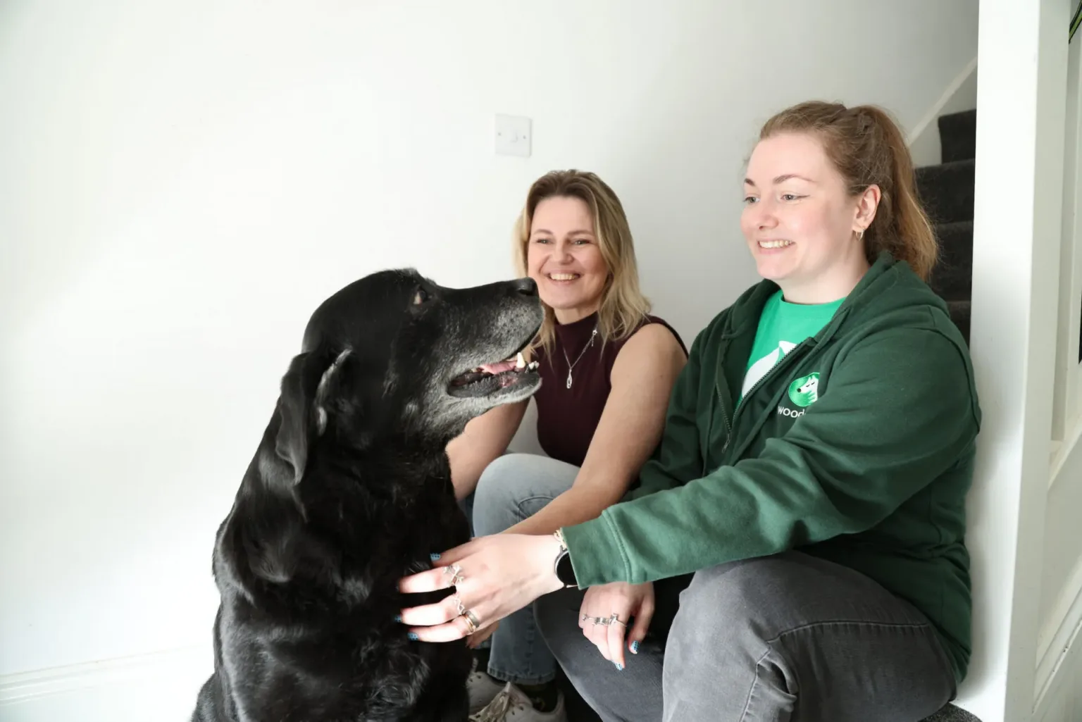 Two women sit and smile at a large black dog indoors by a staircase. One woman wears a green jacket and pets the dog, whilst the other wears a sleeveless top and looks at the dog.
