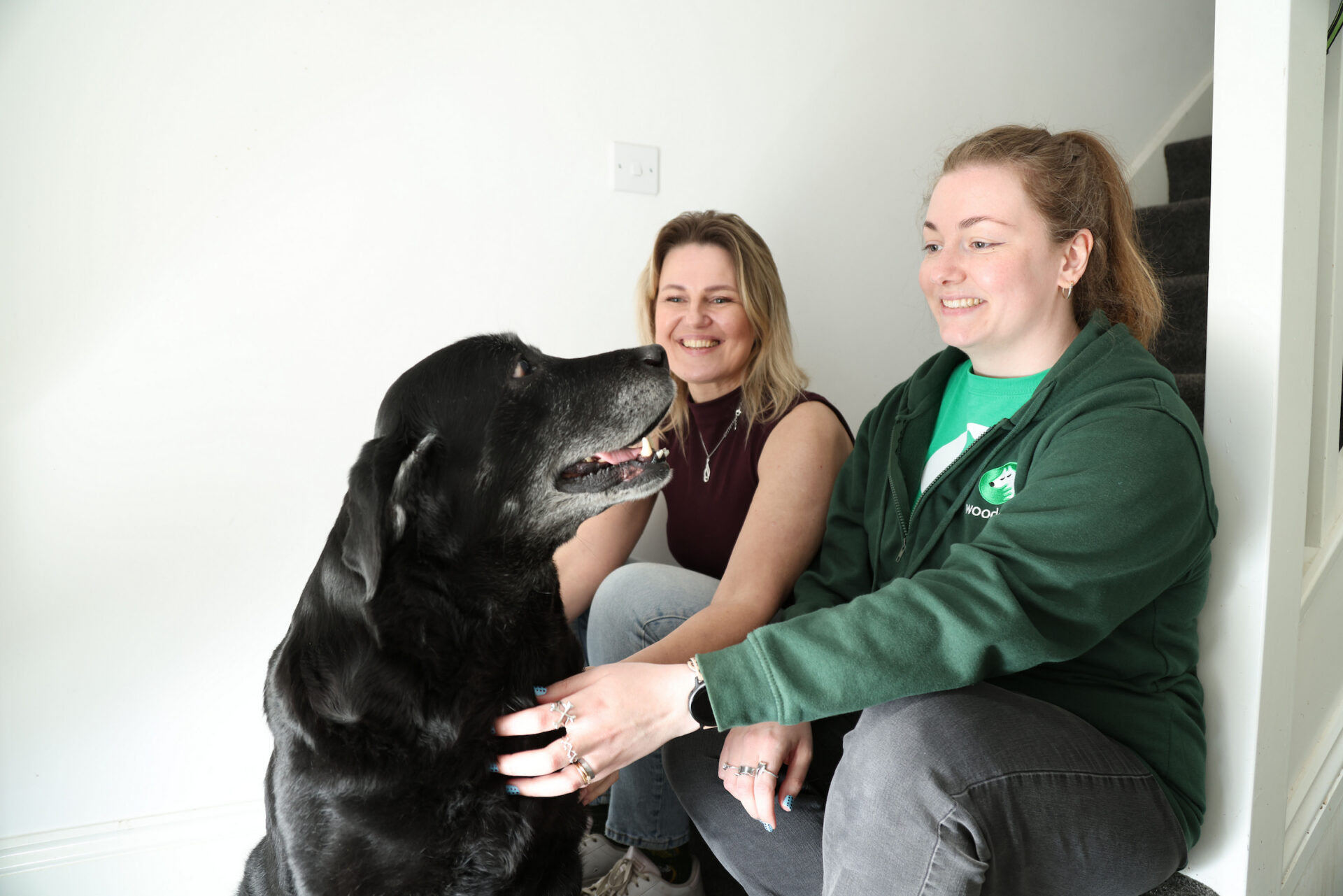 Two women sit and smile at a large black dog indoors by a staircase. One woman wears a green jacket and pets the dog, whilst the other wears a sleeveless top and looks at the dog.